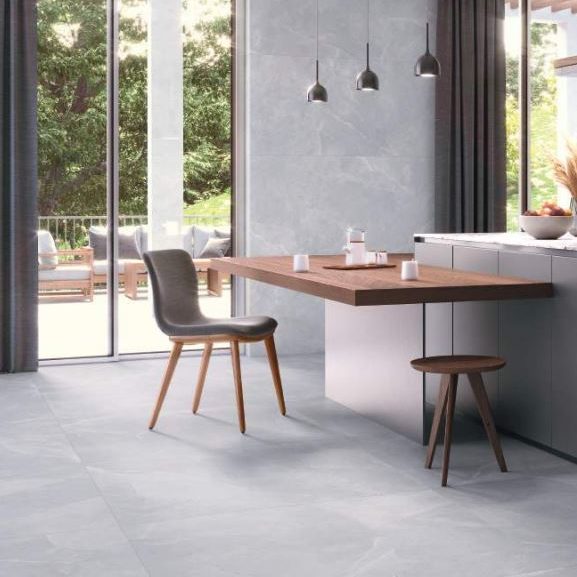 A close-up view of a concrete-like floor tile in a light grey shade, featuring subtle textures and variations. The surface has a matte finish, enhancing the tile's contemporary and industrial aesthetic. A potted plant and a modern chair are partially visible, adding to the minimalist design.