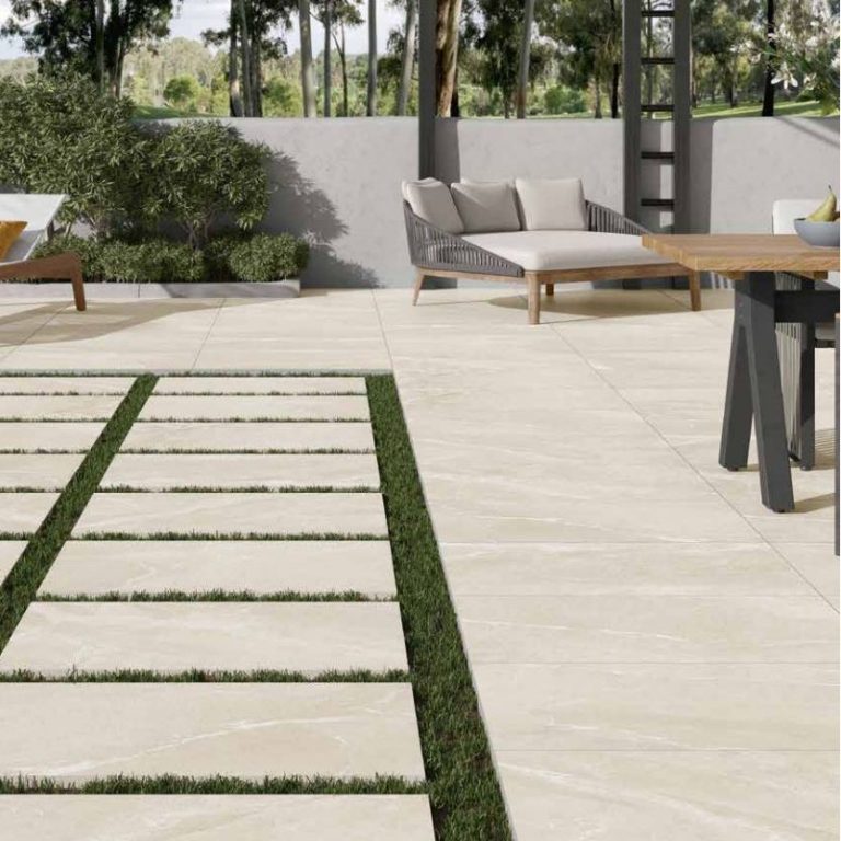 An outdoor patio area with large grey floor tiles that have a natural stone appearance. The space features modern outdoor furniture, including a round dining table with chairs and a lounging area. The clean lines and neutral tones of the tiles complement the sleek architectural elements and lush greenery surrounding the patio.